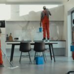 Group of cleaners in red uniforms mopping and wiping glass in a modern dining room.
